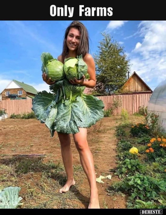 Woman standing barefoot in a garden wearing an outfit made of large cabbages and cabbage leaves, with houses and a greenhouse behind her.