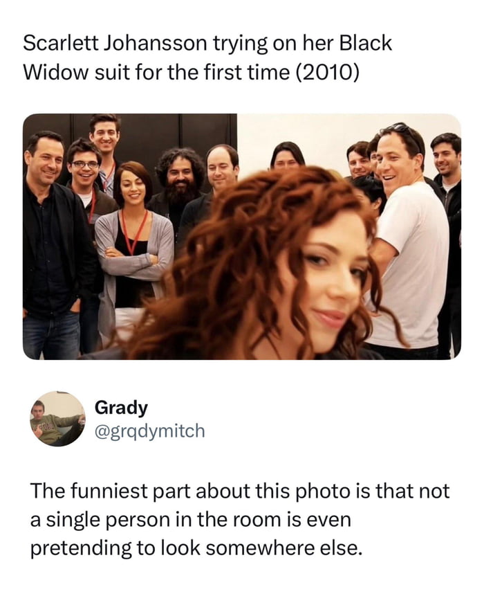 Screenshot of a tweet showing a behind-the-scenes photo of a film crew standing in a room, all looking toward a red-haired woman in the blurred foreground, with a caption joking about the Black Widow suit fitting.